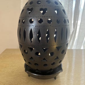 May include: A dark brown, egg-shaped decorative lantern with cut-out heart, circle, and diamond shapes. The lantern sits on a matching round base with three small feet. The design allows light to filter through, creating a warm ambiance.