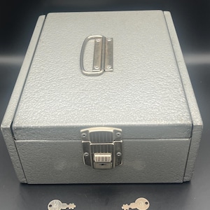May include: A rectangular gray metal lockbox with a textured finish, featuring a silver-colored metal handle on the top and a central locking latch on the front. Two small silver-colored metal keys are positioned in front of the box.
