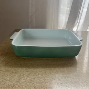 May include: A square baking dish with a light teal interior and a darker teal exterior. The dish has two small handles on either side and is sitting on a light brown wooden surface.