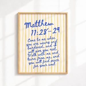 Puede incluir: Una impresión enmarcada con un marco de madera clara. La obra de arte presenta un fondo a rayas amarillas y blancas con texto azul escrito a mano que dice "Mateo 11:28-29" y una cita sobre encontrar descanso y paz.