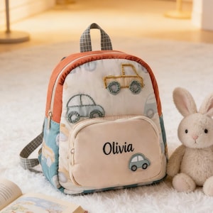 May include: A small, colorful backpack with car appliqués and the name "Olivia" embroidered on the front pocket. A stuffed bunny sits beside the backpack, and an open book is in the foreground. The backpack has a handle and adjustable straps.