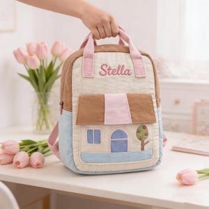May include: A beige, brown, and blue backpack with a house design. The backpack has a pink handle and straps, with the name "Stella" embroidered in pink. The house design includes a blue door and windows, and a tree with red apples.