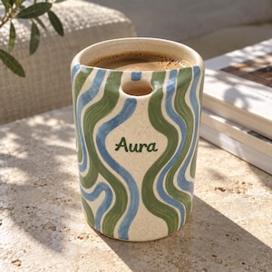 May include: A cylindrical ceramic mug with a speckled cream base featuring hand-painted wavy vertical stripes in shades of sage green and sky blue. The word Aura is printed in green script on the front, and a small circular cutout is near the rim.