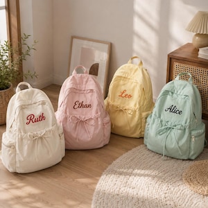 May include: Four backpacks in white, pink, yellow, and light green, each featuring a front drawstring pocket and embroidered names: Ruth, Ethan, Leo, and Alice. The bags have side pockets, top handles, and zipper closures on the main compartments.