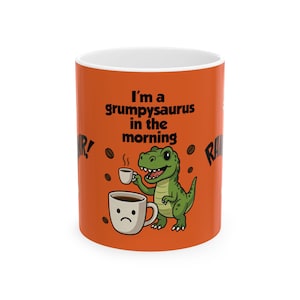 May include: Orange ceramic coffee mug with the text "I'm a grumpy saurus in the morning" and a cartoon dinosaur holding a cup of coffee. The mug also features a sad-faced coffee cup and coffee beans.