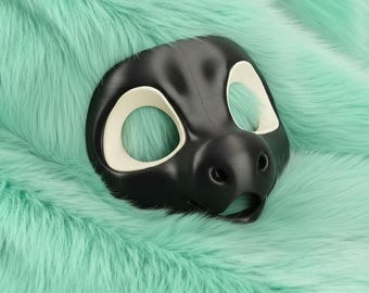 May include: A black and cream-coloured animal mask with large eye openings and a snout. The mask is placed on a bed of soft, mint-green faux fur. The mask is likely for cosplay or costume use.