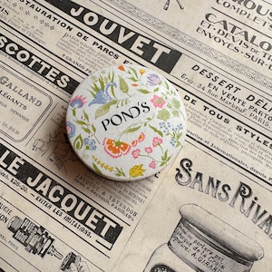 Op de afbeelding: Een ronde, vintage POND'S cosmetica-tin met een bloemenpatroon in roze, oranje, blauw en groen. De tin staat op een achtergrond van oude kranten. Het woord "POND'S" is zwart gedrukt, gecentreerd op het deksel.