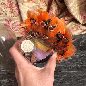 Puede incluir: Un adorno decorativo con un grupo de plumas de color naranja vibrante adornadas con peque&ntilde;as gemas rojas circulares. La base incluye un componente hexagonal blanco y una secci&oacute;n de tela morada, todo cubierto por una c&uacute;pula de cristal.