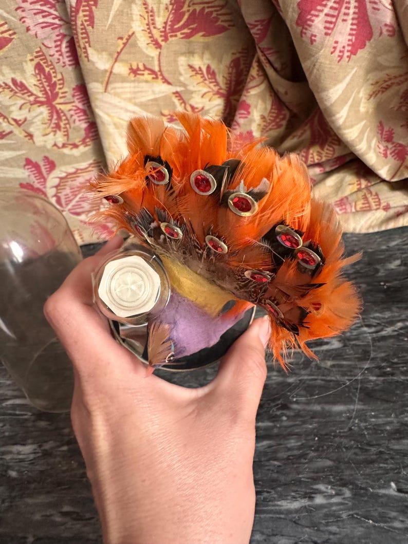 Puede incluir: Un adorno decorativo con un grupo de plumas de color naranja vibrante adornadas con peque&ntilde;as gemas rojas circulares. La base incluye un componente hexagonal blanco y una secci&oacute;n de tela morada, todo cubierto por una c&uacute;pula de cristal.