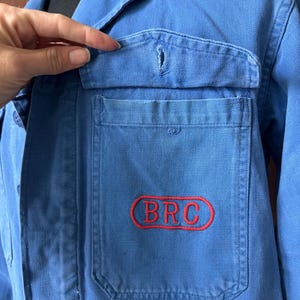 May include: A blue work jacket featuring a collared neckline, long sleeves, and a front chest pocket with a flap. The pocket is embroidered with the red text BRC enclosed within a red oval outline. The jacket is constructed from a durable fabric.