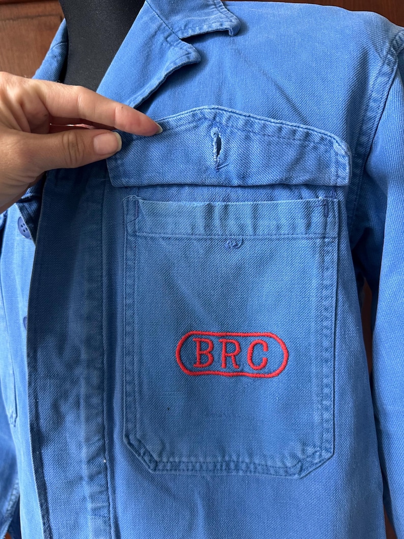 May include: A blue work jacket featuring a collared neckline, long sleeves, and a front chest pocket with a flap. The pocket is embroidered with the red text BRC enclosed within a red oval outline. The jacket is constructed from a durable fabric.