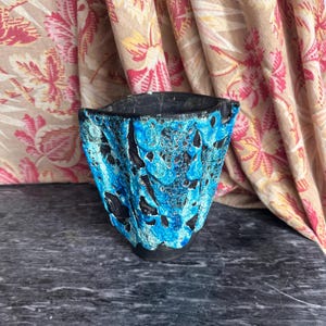 May include: A ceramic vessel featuring a dark black base with a thick, textured, and cratered bright blue glaze applied to the exterior. The object has an irregular, organic shape with a wide, uneven rim and a tapered bottom base.