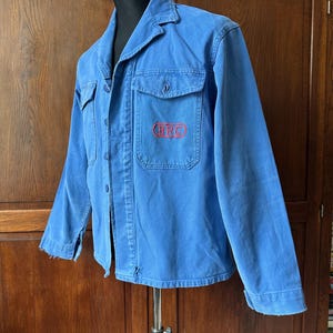May include: A blue long-sleeve work jacket featuring a collared neckline, a button-down front, and two chest pockets. The left chest pocket displays the red embroidered text BRC inside an oval outline. The jacket has visible wear at the hem.