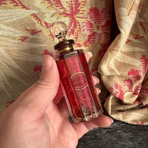 Puede incluir: Un frasco de perfume rojo vintage con un tapón de cristal transparente. La botella tiene detalles dorados y una etiqueta con texto. La botella es rectangular con bordes redondeados. El fondo presenta una tela con estampado floral.