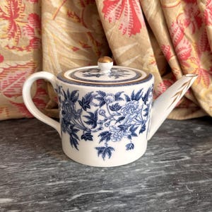 Op de afbeelding: Een witte keramische theepot met een blauw bloemenpatroon van pioenrozen en bladeren. De theepot heeft een cilindervormig lichaam, een gebogen handvat, een rechte tuit met gouden accenten en een bijpassend deksel met een kleine gouden knop.