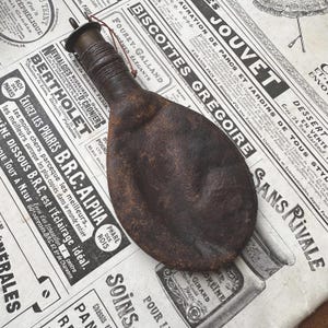 Peut inclure: Une gourde en cuir brun foncé, plate et circulaire, dotée d'un goulot cylindrique étroit entouré d'un cordon en cuir. La gourde présente un petit bouchon métallique au sommet et une fine ficelle rouge foncé attachée au goulot, à la texture usée.