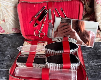 Puede incluir: Un kit de aseo de viaje rojo con cremallera que contiene varias herramientas, incluyendo un espejo rectangular, dos cepillos de cerdas blancas y negras, un tubo de plástico transparente, un calzador de metal, tijeras pequeñas y utensilios de manicura.