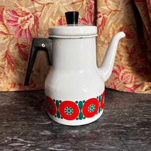 Könnte beinhalten: Eine weiße Emaille-Kaffeekanne mit schwarzem Griff und schwarzem Knauf auf dem Deckel. Der Körper der Kanne ist im unteren Bereich mit einem sich wiederholenden horizontalen Muster aus großen roten Kreisen mit grünen Sternen verziert.