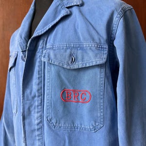 May include: A light blue work jacket featuring a collared neckline, long sleeves, and two front chest pockets. The left chest pocket displays the letters BRC embroidered in red within a red oval outline. The jacket is made of a durable cotton fabric.