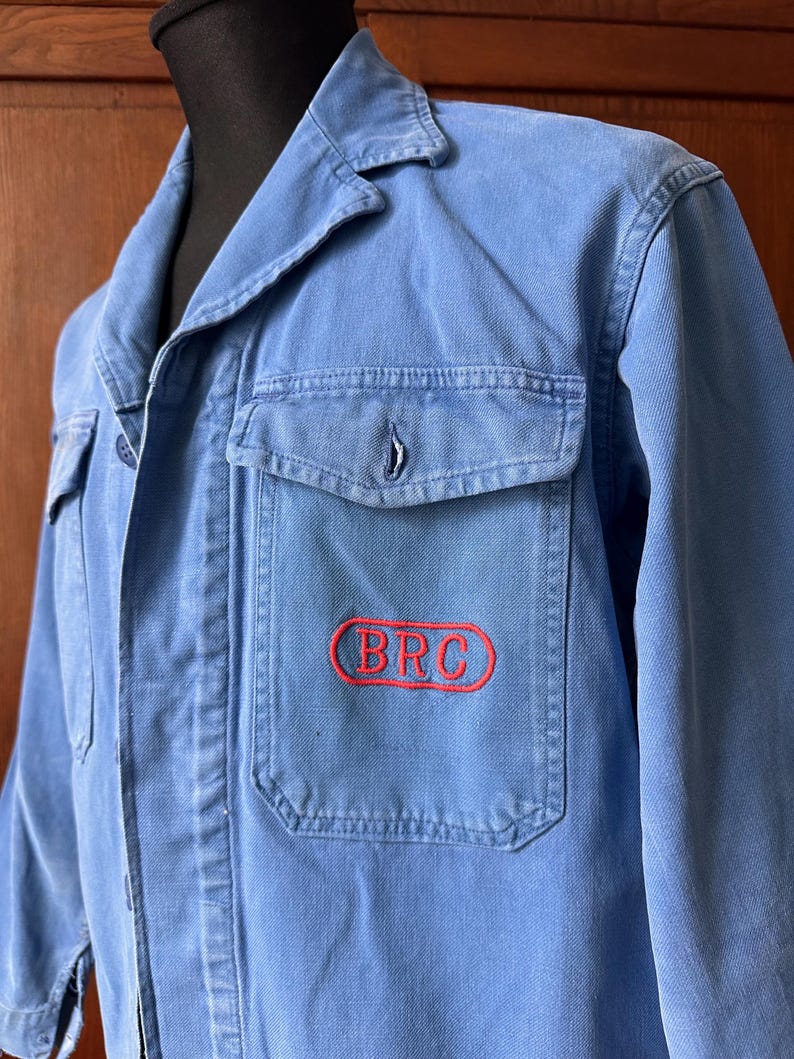 May include: A light blue work jacket featuring a collared neckline, long sleeves, and two front chest pockets. The left chest pocket displays the letters BRC embroidered in red within a red oval outline. The jacket is made of a durable cotton fabric.