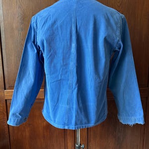 May include: A bright blue long-sleeved jacket featuring a classic collar, a vertical seam running down the center of the back, and buttoned cuffs. The garment is displayed on a mannequin, showcasing a solid color design with visible stitching.
