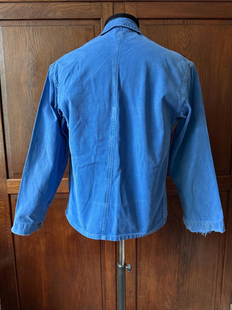May include: A bright blue long-sleeved jacket featuring a classic collar, a vertical seam running down the center of the back, and buttoned cuffs. The garment is displayed on a mannequin, showcasing a solid color design with visible stitching.