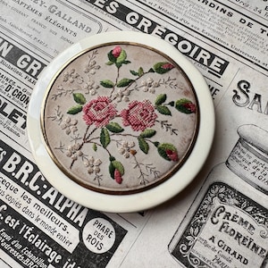 Puede incluir: Un compacto vintage de color marfil con un diseño de bordado floral detallado. El bordado presenta rosas rosas, hojas verdes y pequeñas flores blancas. El compacto tiene un borde de metal dorado y está sobre un fondo de recortes de periódicos antiguos.