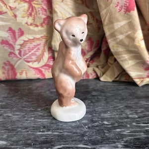 Peut inclure: Petite figurine vintage en céramique représentant un ours en peluche. L'ours est debout sur une base blanche, avec un corps brun clair et une tête rose clair. Il a de petits yeux noirs et un nez noir. Un charmant objet décoratif.