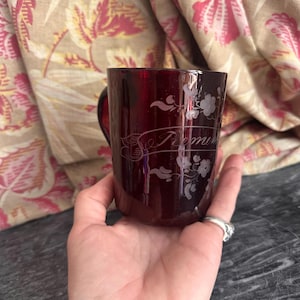 Pode incluir: Uma caneca de vidro vermelho profundo com um design floral gravado e uma faixa com a palavra "Remember" inscrita na frente. A caneca possui uma superfície lisa e translúcida, uma alça curva e uma forma cilíndrica clássica de vidro.