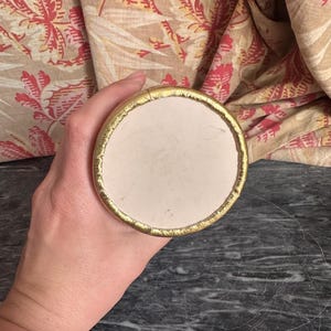 Puede incluir: Un objeto circular con una superficie central lisa de color blanco roto y un borde met&aacute;lico dorado texturizado. El objeto se sostiene de manera que muestra la parte superior plana y redonda, resaltando el contraste entre el interior mate.