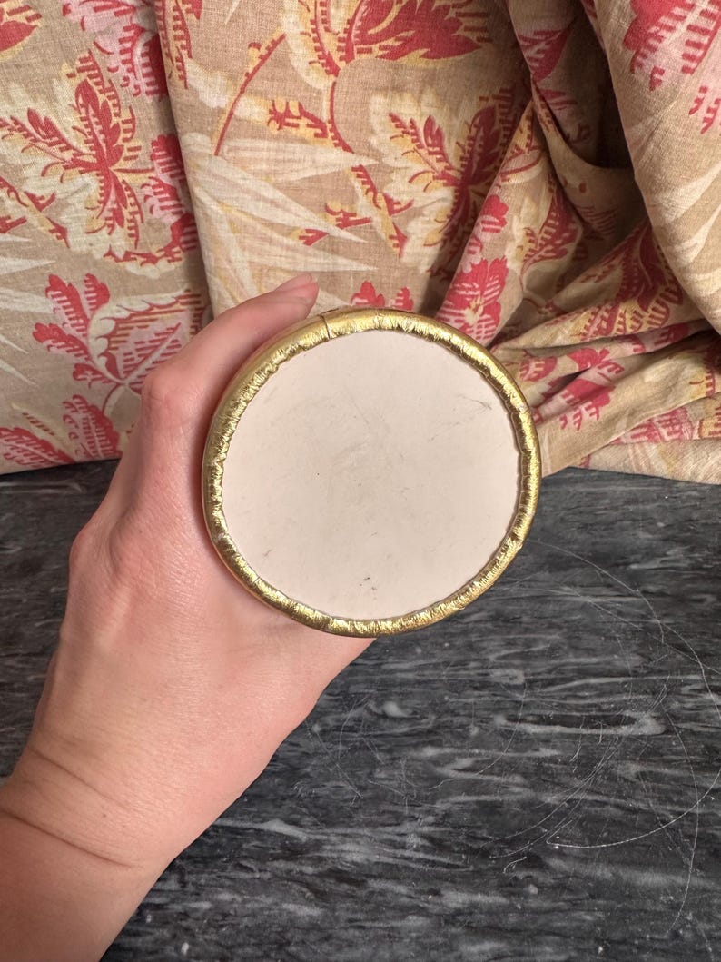 Puede incluir: Un objeto circular con una superficie central lisa de color blanco roto y un borde met&aacute;lico dorado texturizado. El objeto se sostiene de manera que muestra la parte superior plana y redonda, resaltando el contraste entre el interior mate.