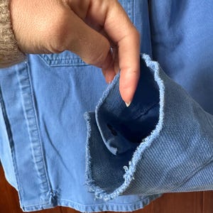 May include: A light blue denim jacket featuring a frayed, raw-edge cuff detail. The jacket is constructed from a sturdy, woven cotton fabric with visible stitching along the seams and a single dark button visible inside the sleeve opening.