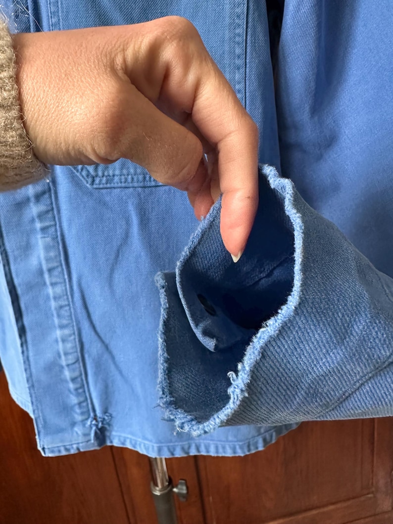 May include: A light blue denim jacket featuring a frayed, raw-edge cuff detail. The jacket is constructed from a sturdy, woven cotton fabric with visible stitching along the seams and a single dark button visible inside the sleeve opening.