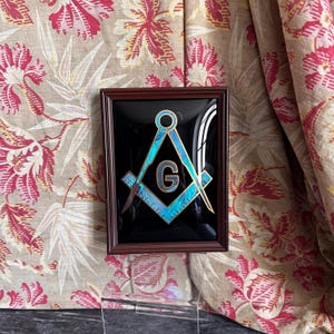 Pode incluir: Uma peça de arte de parede retangular emoldurada com um fundo preto e o emblema maçónico de esquadro e compasso ao centro. O emblema apresenta um acabamento azul iridescente com uma letra G dourada no centro, dentro de uma moldura de madeira escura.