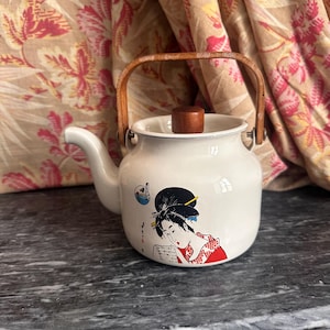 Peut inclure: Théière en émail blanc avec une poignée et un couvercle en bois. La théière présente une illustration de geisha japonaise en rouge, noir et blanc. Le bec verseur est blanc et la poignée est en bois clair. Un article de cuisine décoratif.