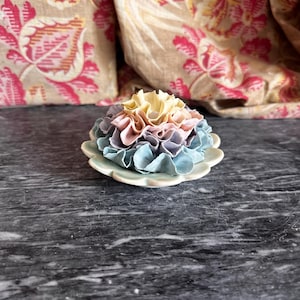 Puede incluir: Un objeto decorativo de cerámica con un diseño de flor de múltiples capas con volantes. Los pétalos transicionan de un amarillo pálido en el centro a rosa suave, lavanda y azul claro en la base, sobre un plato circular de bordes festoneados.
