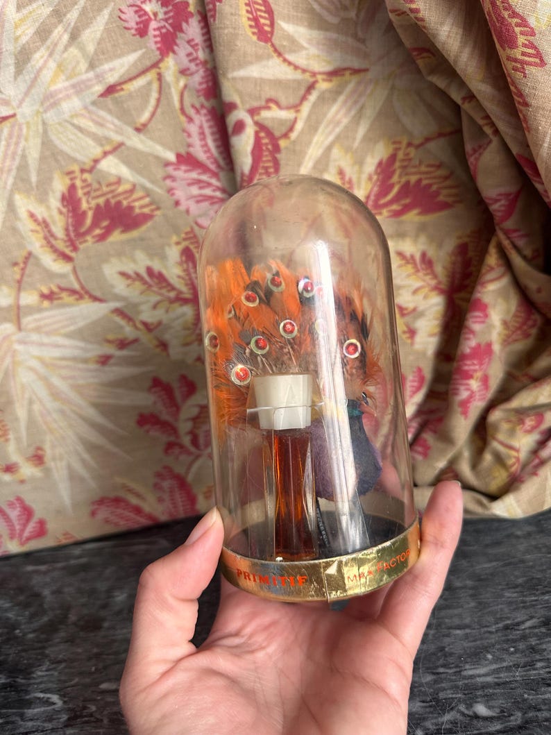 Puede incluir: Una c&uacute;pula de cristal transparente que contiene un peque&ntilde;o frasco de perfume con tap&oacute;n blanco y un arreglo decorativo de plumas naranjas acentuadas con peque&ntilde;os detalles circulares rojos. La base es dorada con el texto PRIMITIF MAX FACTOR.