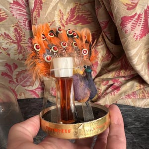 Puede incluir: Un frasco de perfume vintage Primitif Max Factor con un adorno de pavo real de plumas naranjas y lentejuelas rojas. El frasco de cristal transparente contiene l&iacute;quido &aacute;mbar y se asienta sobre una base circular dorada con texto.