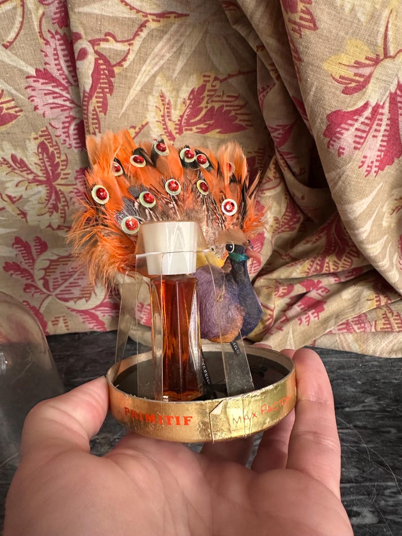 Puede incluir: Un frasco de perfume vintage Primitif Max Factor con un adorno de pavo real de plumas naranjas y lentejuelas rojas. El frasco de cristal transparente contiene l&iacute;quido &aacute;mbar y se asienta sobre una base circular dorada con texto.