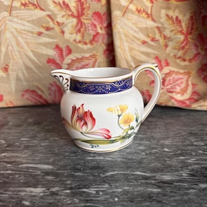 Puede incluir: Una jarrita de leche de porcelana blanca con una banda decorativa azul oscuro y borde dorado cerca de la abertura. El cuerpo presenta un colorido patrón floral con tulipanes rojos y amarillos y hojas verdes, junto a un asa y pico curvos.