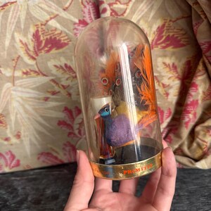 Puede incluir: Una c&uacute;pula de cristal transparente que contiene una peque&ntilde;a figura de p&aacute;jaro morada y naranja con plumas decorativas. La base de la c&uacute;pula presenta una banda met&aacute;lica dorada con el texto Max Factor y Primitif impreso en letras negras.