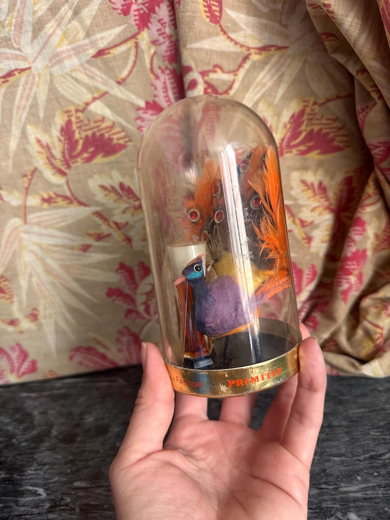 Puede incluir: Una c&uacute;pula de cristal transparente que contiene una peque&ntilde;a figura de p&aacute;jaro morada y naranja con plumas decorativas. La base de la c&uacute;pula presenta una banda met&aacute;lica dorada con el texto Max Factor y Primitif impreso en letras negras.