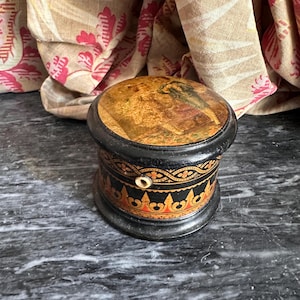 May include: A small, round, antique wooden box with a lid. The box is painted black with gold and red decorative accents. The lid features a painted scene with a gold and brown background. A small hole is visible on the side.