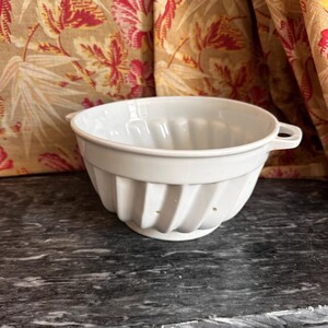 May include: A white ceramic bowl featuring a fluted, ribbed exterior design and two small, integrated side handles. The bowl has a smooth, glossy finish and a circular base, with small drainage holes visible along the lower fluted sections.