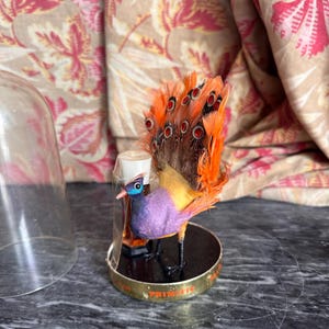 Puede incluir: Una figura decorativa de ave con pecho morado, cuerpo amarillo y plumas de cola de color naranja vibrante con manchas oscuras en forma de ojo. El ave se asienta sobre una base circular negra con borde dorado marcada PRIMITIF, junto a un frasco.