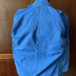 May include: A bright blue denim jacket shown from the back, featuring a structured collar, a vertical center seam, and long sleeves. The fabric has a slightly faded appearance with visible stitching details along the seams and a frayed hemline.