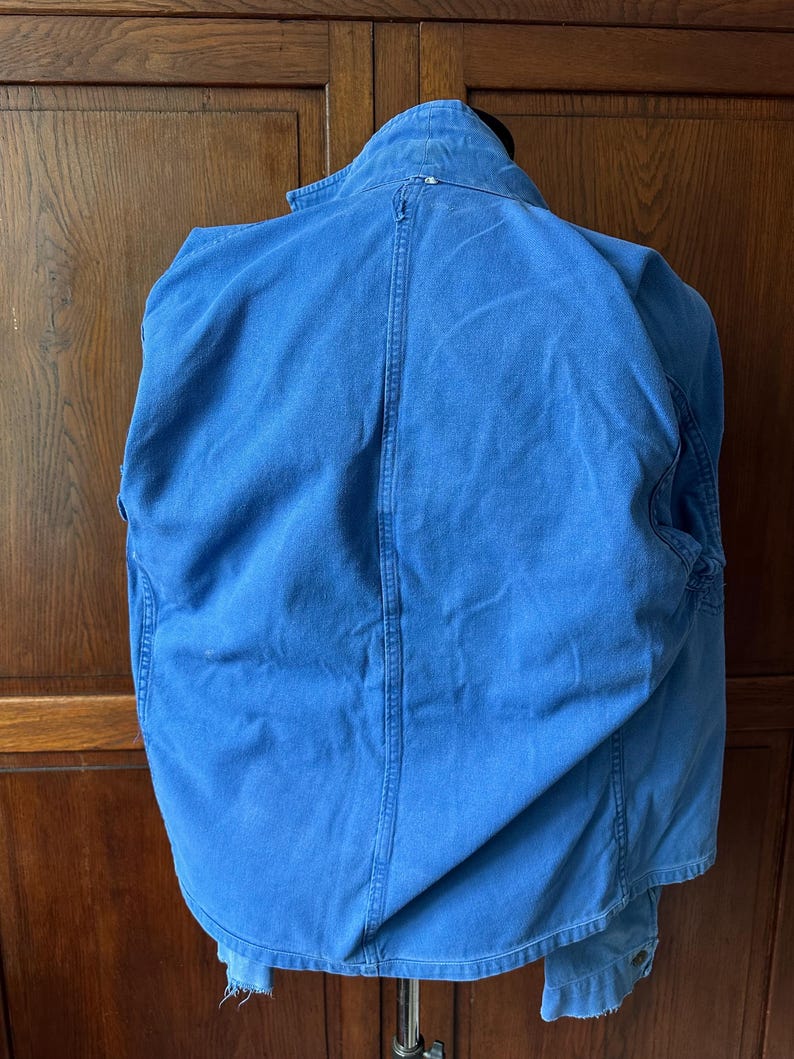 May include: A bright blue denim jacket shown from the back, featuring a structured collar, a vertical center seam, and long sleeves. The fabric has a slightly faded appearance with visible stitching details along the seams and a frayed hemline.