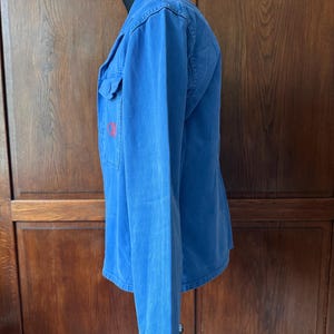 May include: A long-sleeved blue button-down shirt featuring a classic collar and a single chest pocket on the left side. The garment displays a small, circular red embroidered logo on the pocket area and is shown from a side profile view.