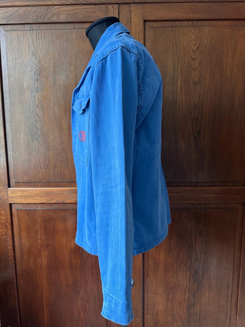May include: A long-sleeved blue button-down shirt featuring a classic collar and a single chest pocket on the left side. The garment displays a small, circular red embroidered logo on the pocket area and is shown from a side profile view.