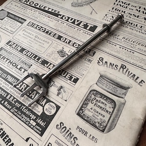 Peut inclure: Un outil argenté de style vintage avec une extrémité en forme de fourche et une longue poignée, posé sur un fond de journal. Un pot vintage avec le texte "Crème Floréine" est également visible.